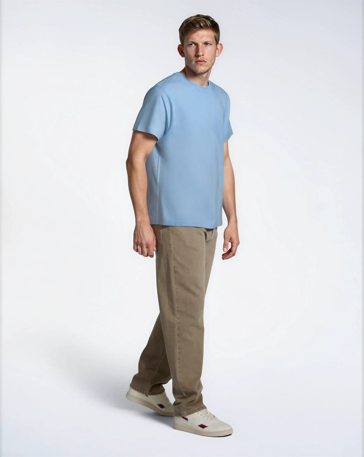 A full body photo of a man looking away from the camera, modelling a plain blue, short sleeve organic cotton t shirt with brown trousers.