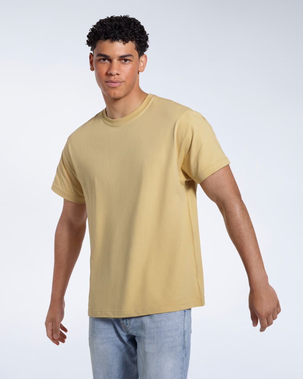 A man looking into the camera with his arms slightly raised, modelling a plain light khaki short sleeve organic cotton t shirt.