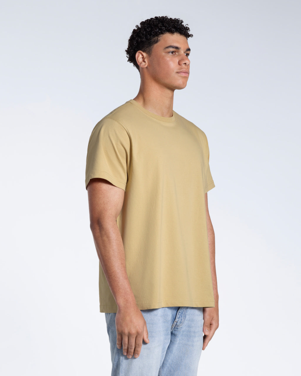 A man at an angle with his arms by his sides, modelling a plain light khaki short sleeve organic cotton t shirt.