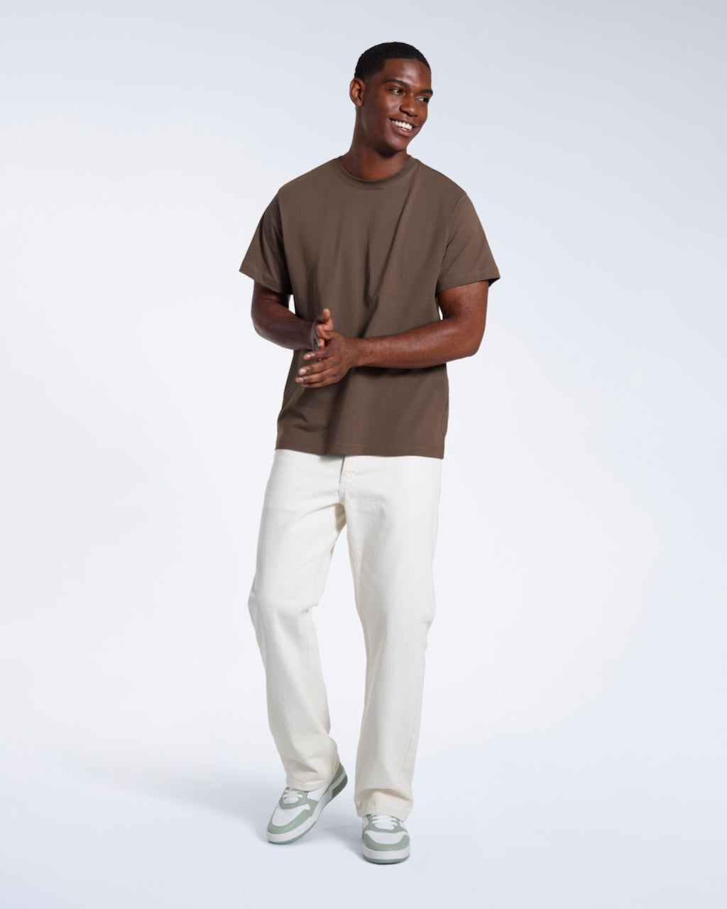 A full body photo of a man smiling with his hands together, modelling a plain dark khaki short sleeve organic cotton t shirt.