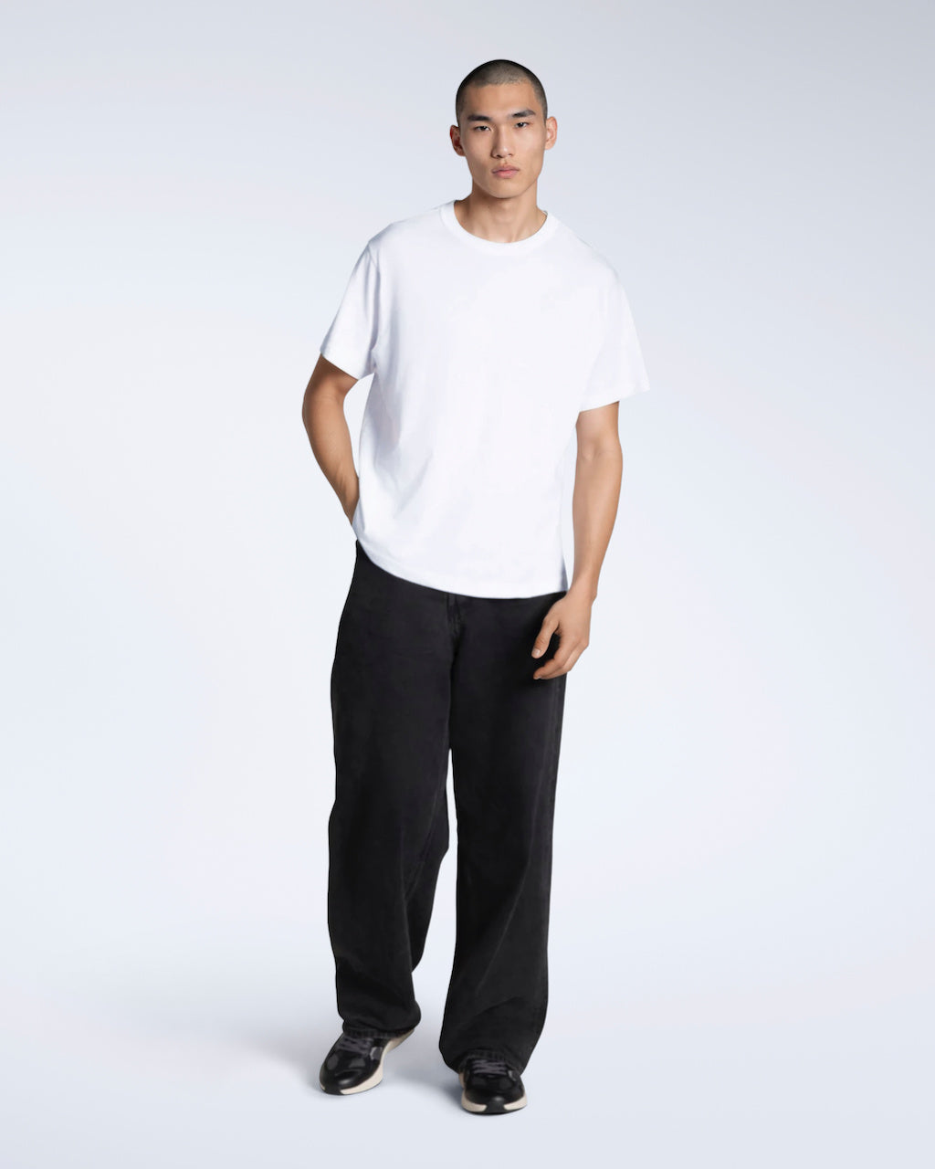 A full body photo of a man looking into the camera with his hand in his pocket, modelling a plain white, short sleeve organic cotton t shirt with a pair of jeans.