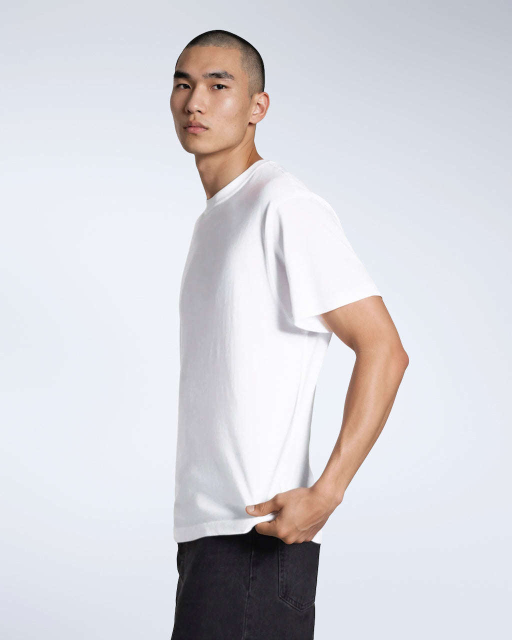 A man facing side on looking into the camera, modelling a plain white, short sleeve organic cotton t shirt.