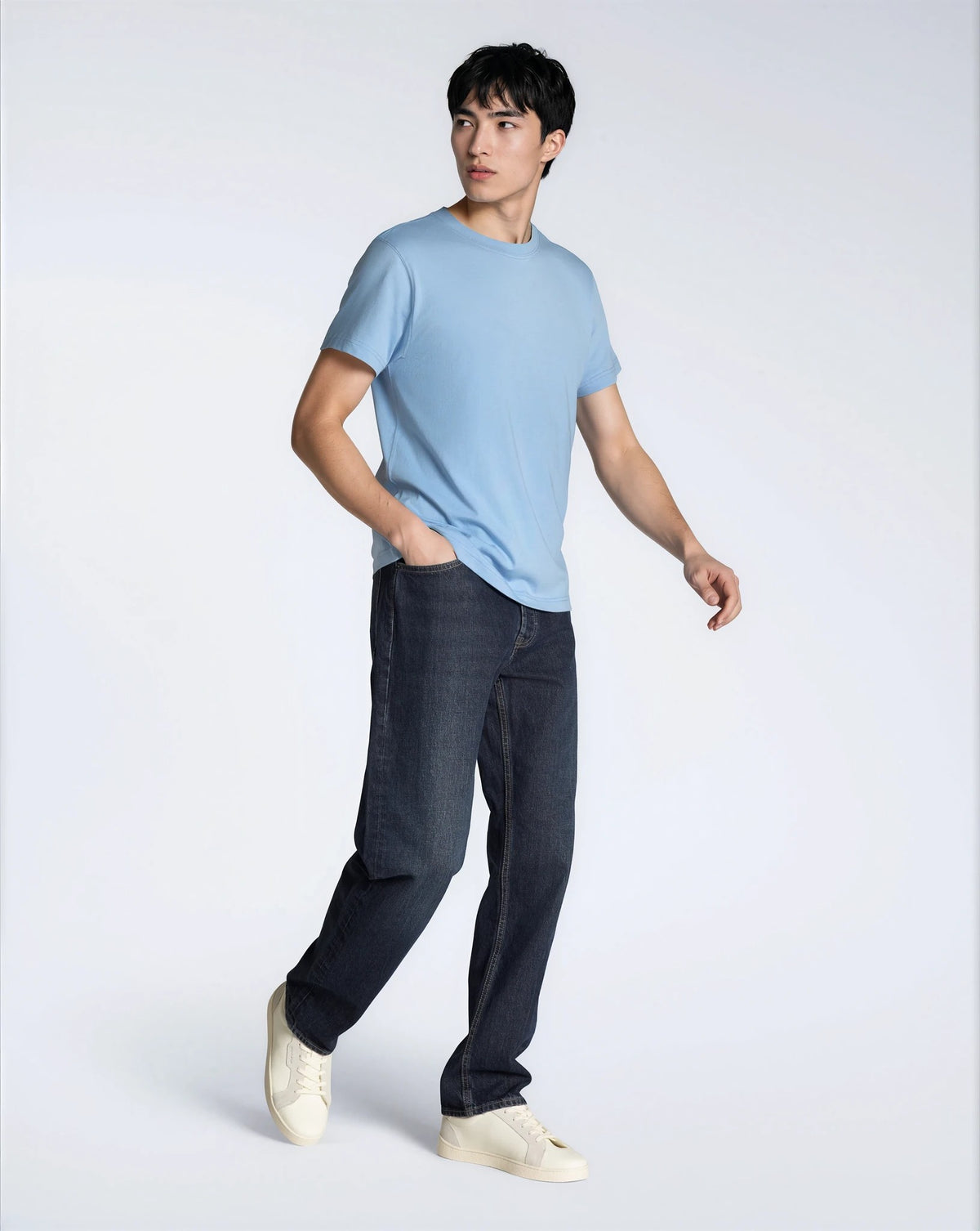 A full body photo of a man looking away from the camera with his hand in his pocket, modelling a plain blue, short sleeve organic cotton t shirt with a pair of jeans.