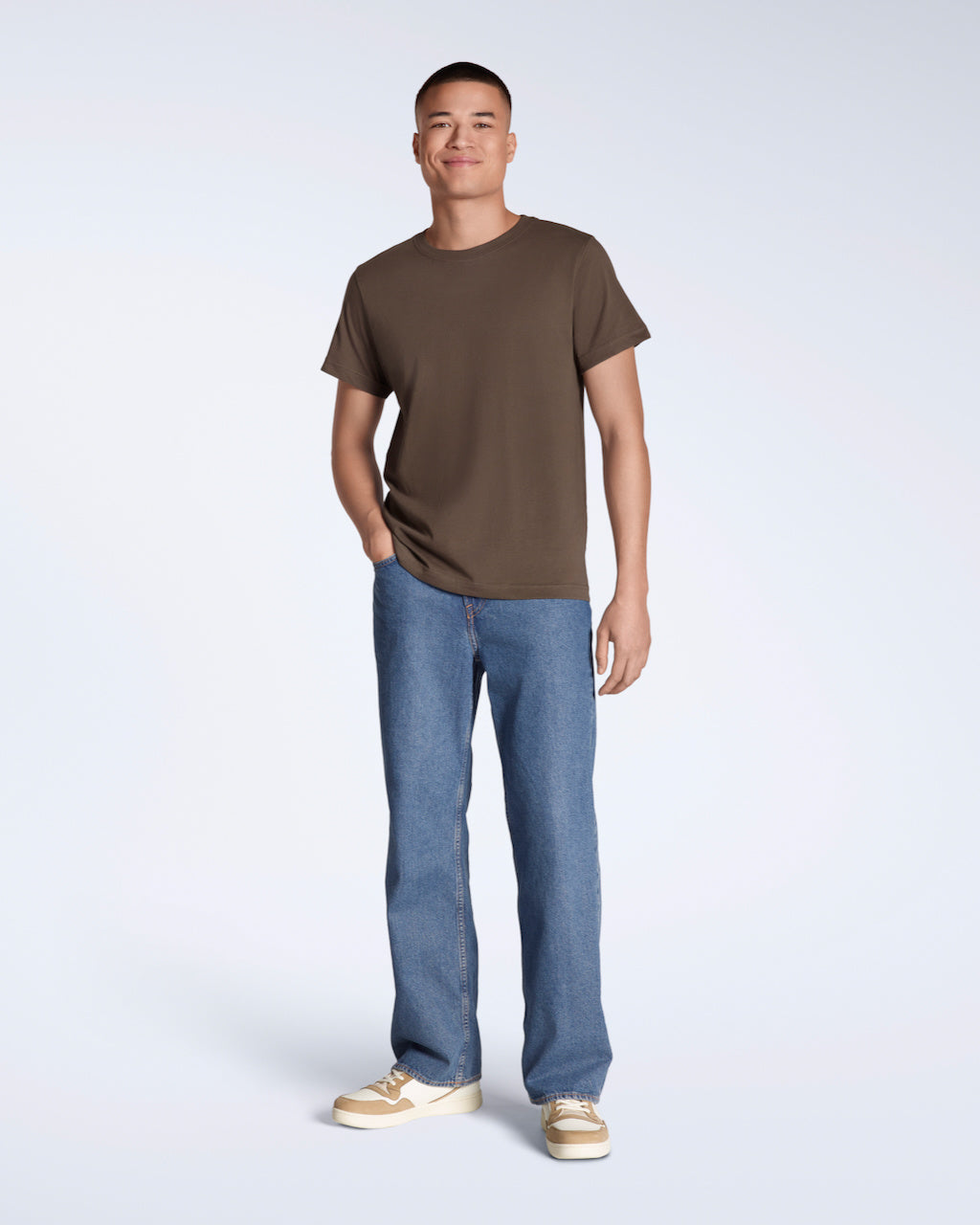 A full body photo of a man smiling into the camera with his hand in his pocket, modelling a plain brown, short sleeve organic cotton t shirt with a pair of jeans.