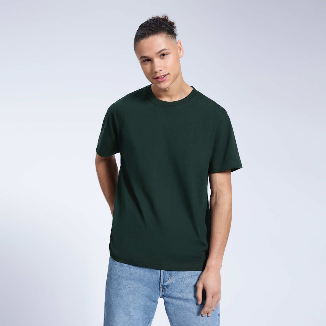 A man smiling into the camera with one hand in his back pocket, modelling a plain green short sleeve organic cotton t shirt.