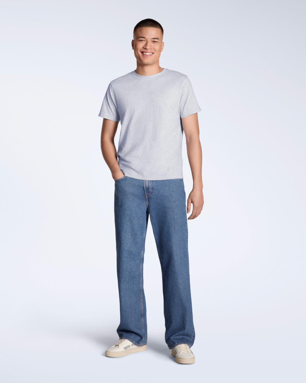 A full body photo of a man smiling into the camera with his hand in his pocket, modelling a plain grey short sleeve organic cotton t shirt with a pair of jeans.