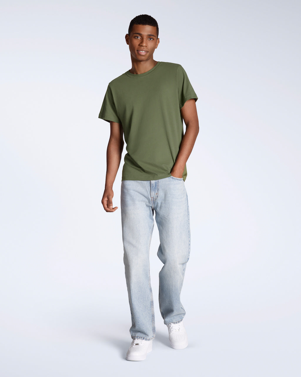 A full body photo of a man looking into the camera with his hand in his pocket, modelling a plain khaki green, short sleeve organic cotton t shirt with a pair of jeans.