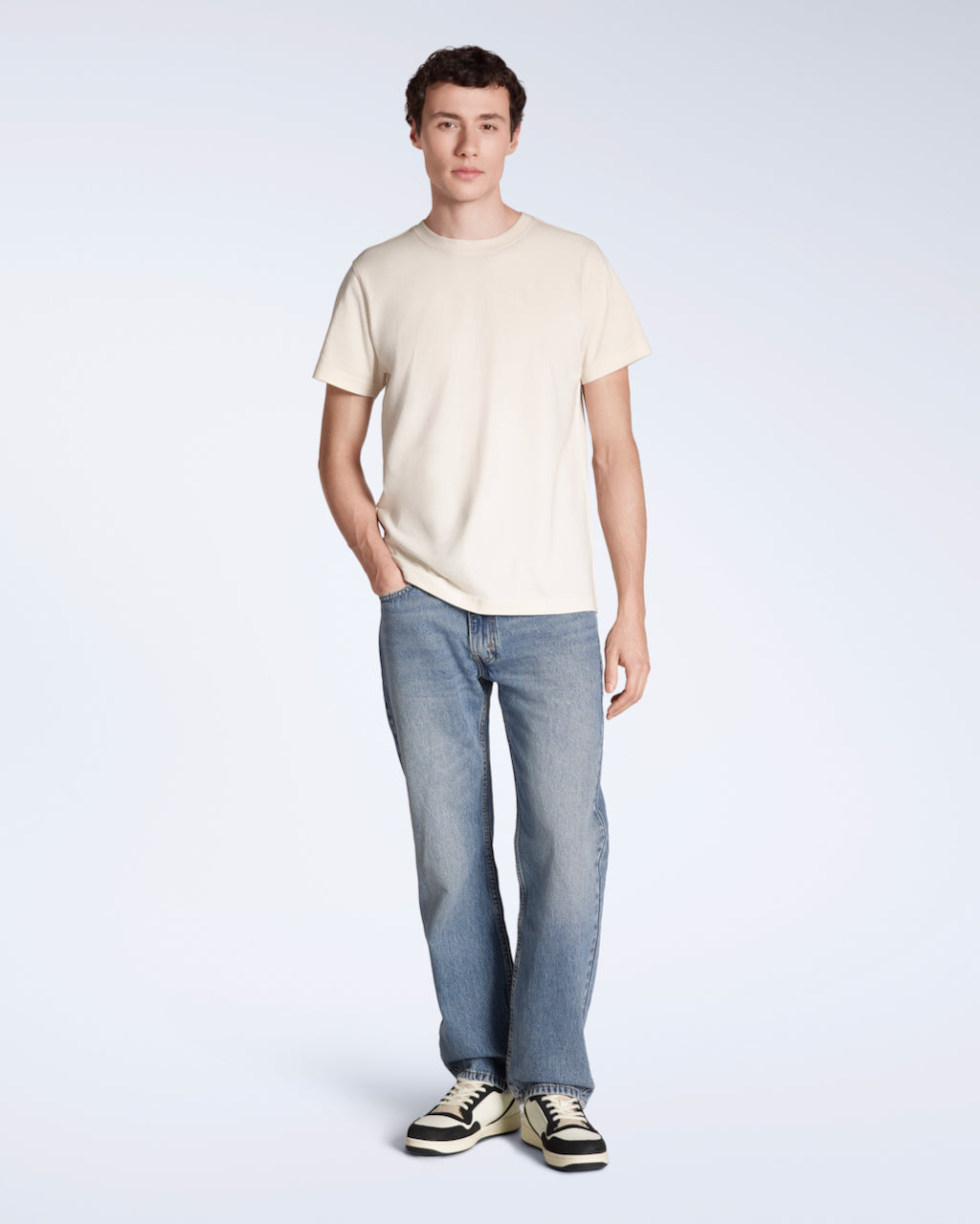 A full body photo of a man looking into the camera with his hand in his pocket, modelling a plain natural coloured, short sleeve organic cotton t shirt with a pair of jeans.