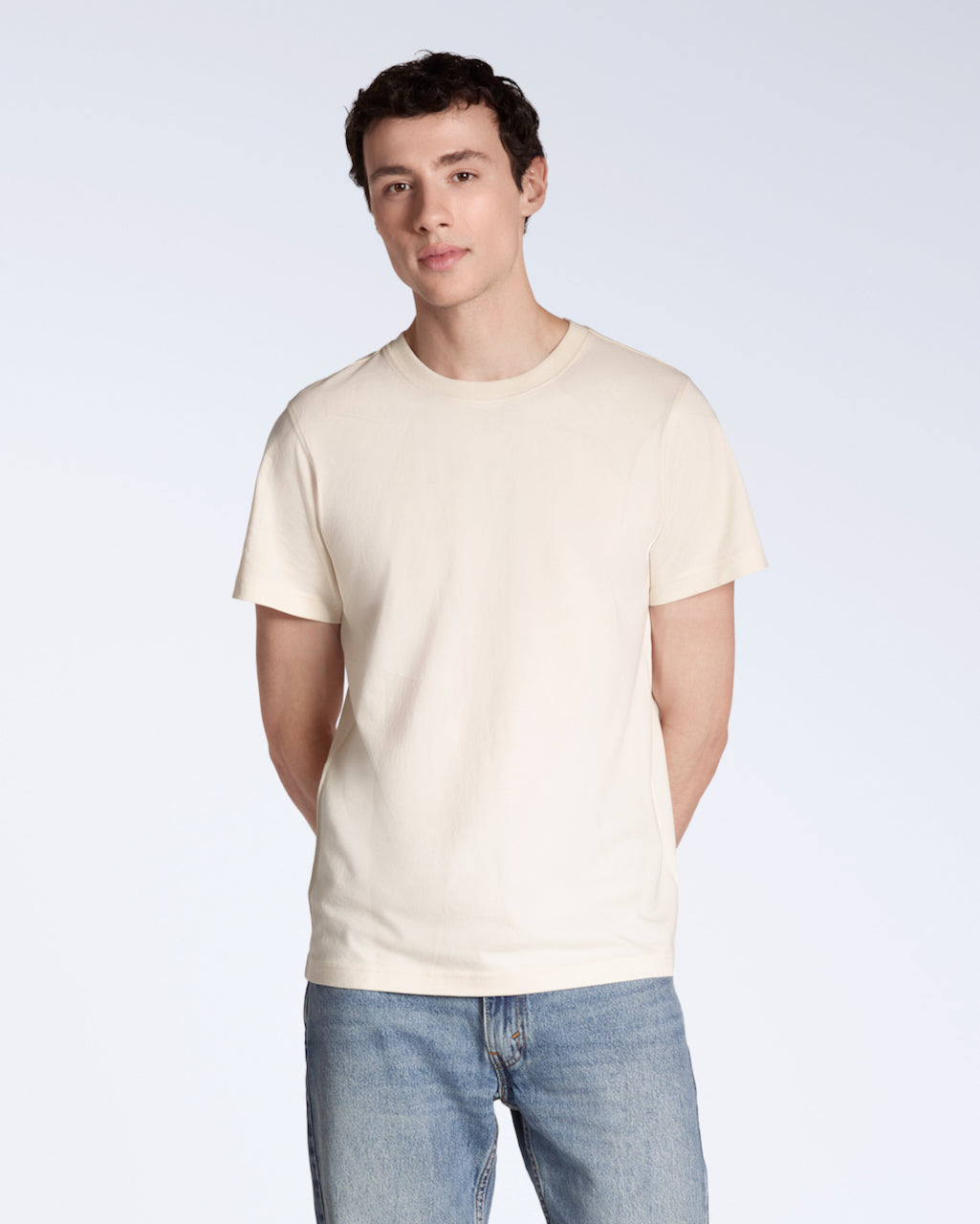 A man looking into the camera with his hands behind his back, modelling a plain natural coloured, organic cotton t shirt.