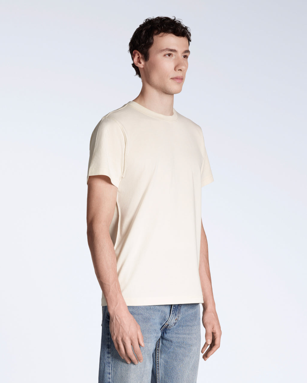 A man at an angle with his arms by his sides, modelling a plain natural coloured short sleeve organic cotton t shirt.