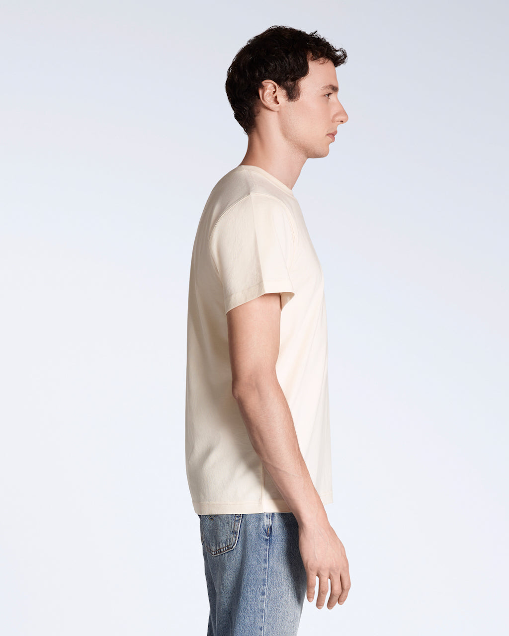 A man facing side on, modelling a plain natural coloured, short sleeve, organic cotton t shirt.