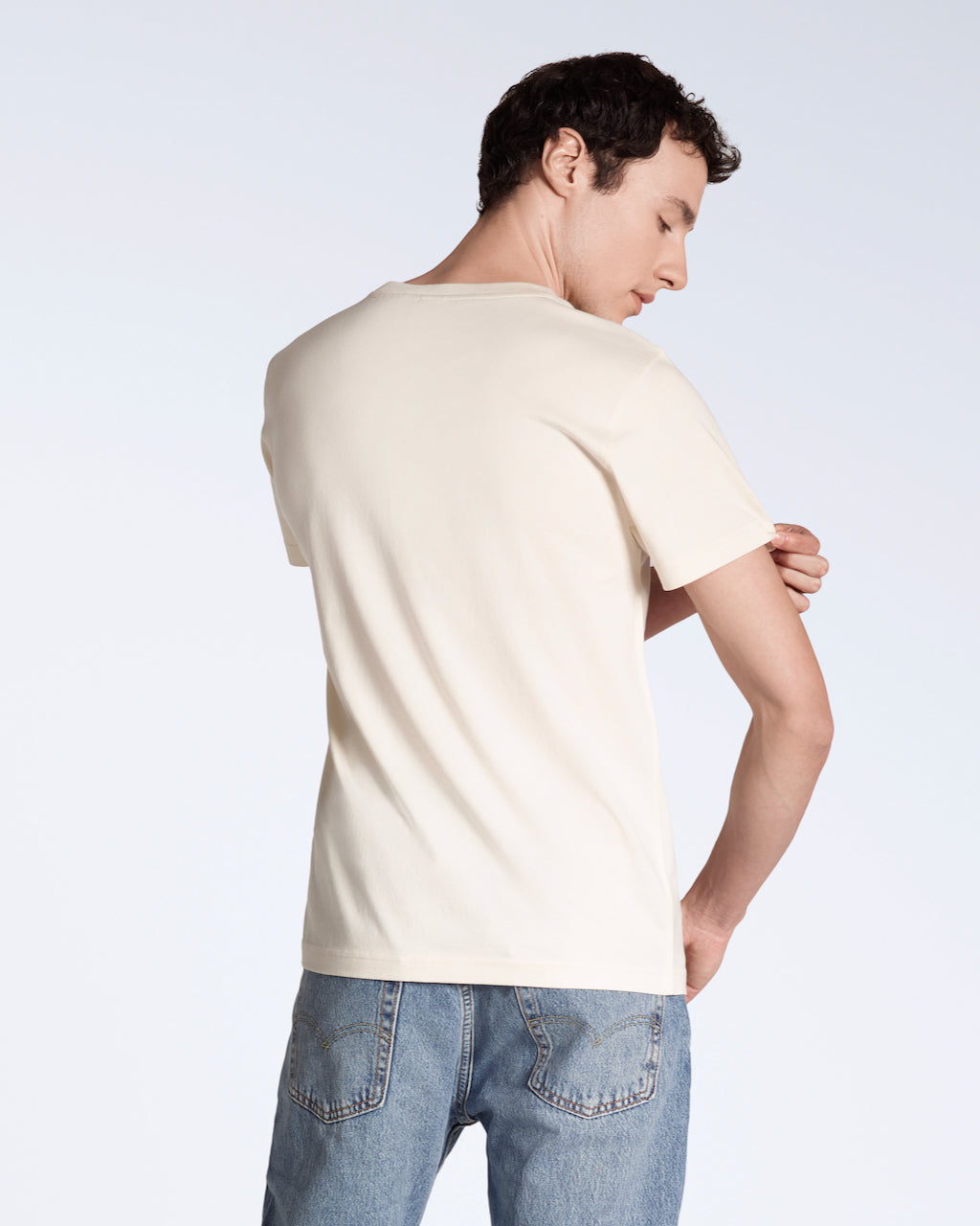 A man facing away from the camera touching his sleeve, modelling a plain natural coloured, short sleeve, organic cotton t shirt.