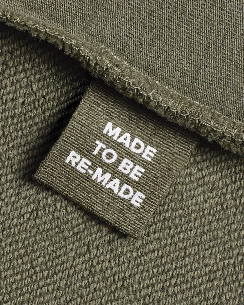 A close up of a label tag in a olive, organic cotton overshirt that says 'made to be re made'.