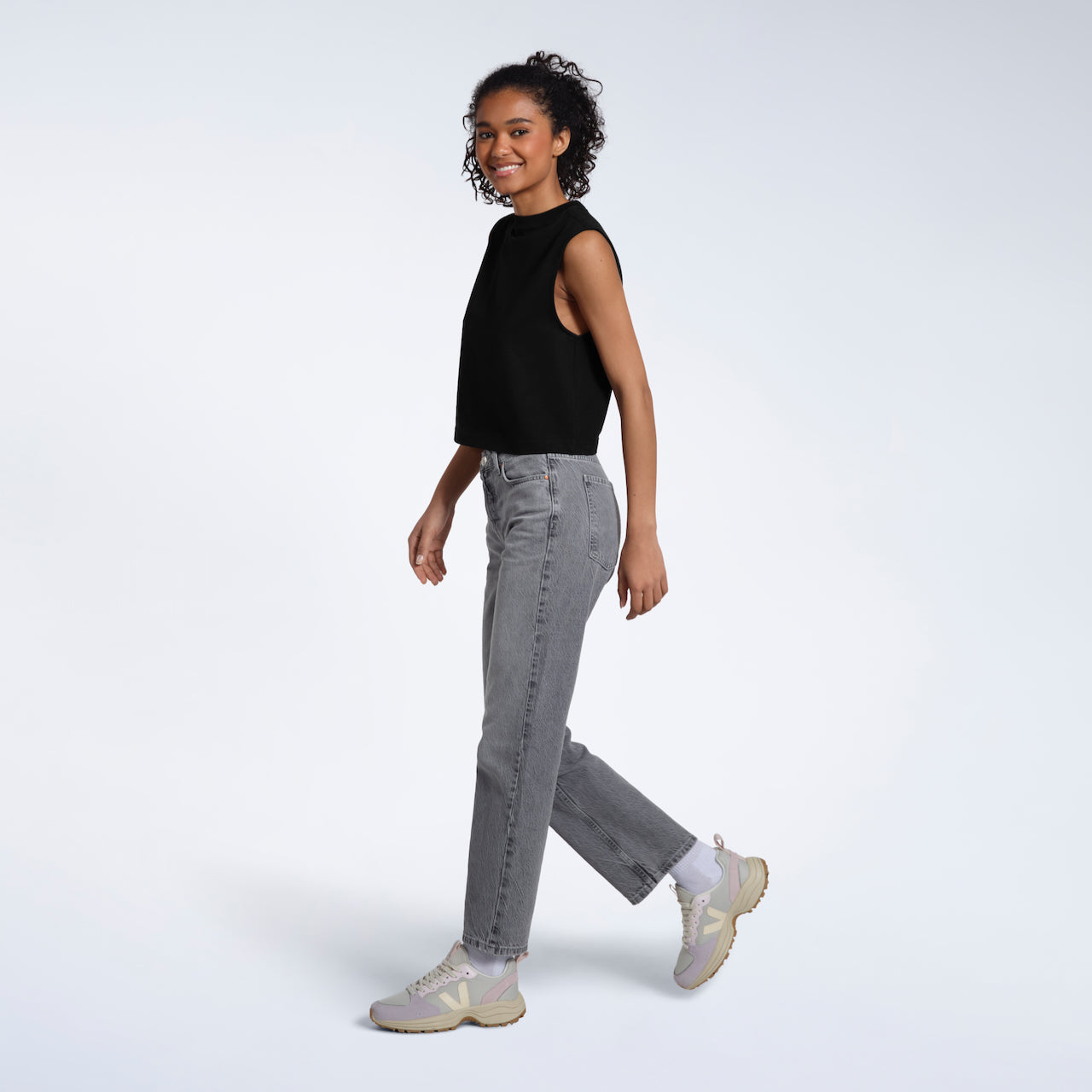 A full body photo of a woman walking and smiling, modelling a plain black sleeveless organic cotton tank top.