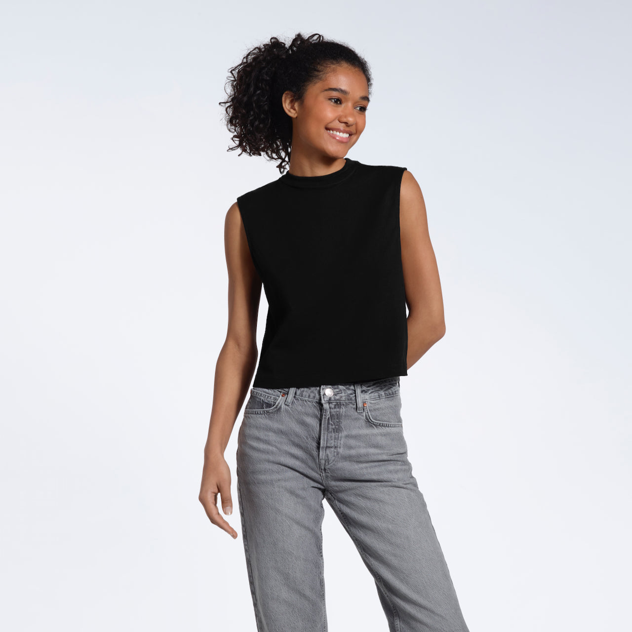 A woman smiling with one arm behind her, modelling a plain black sleeveless organic cotton tank top.