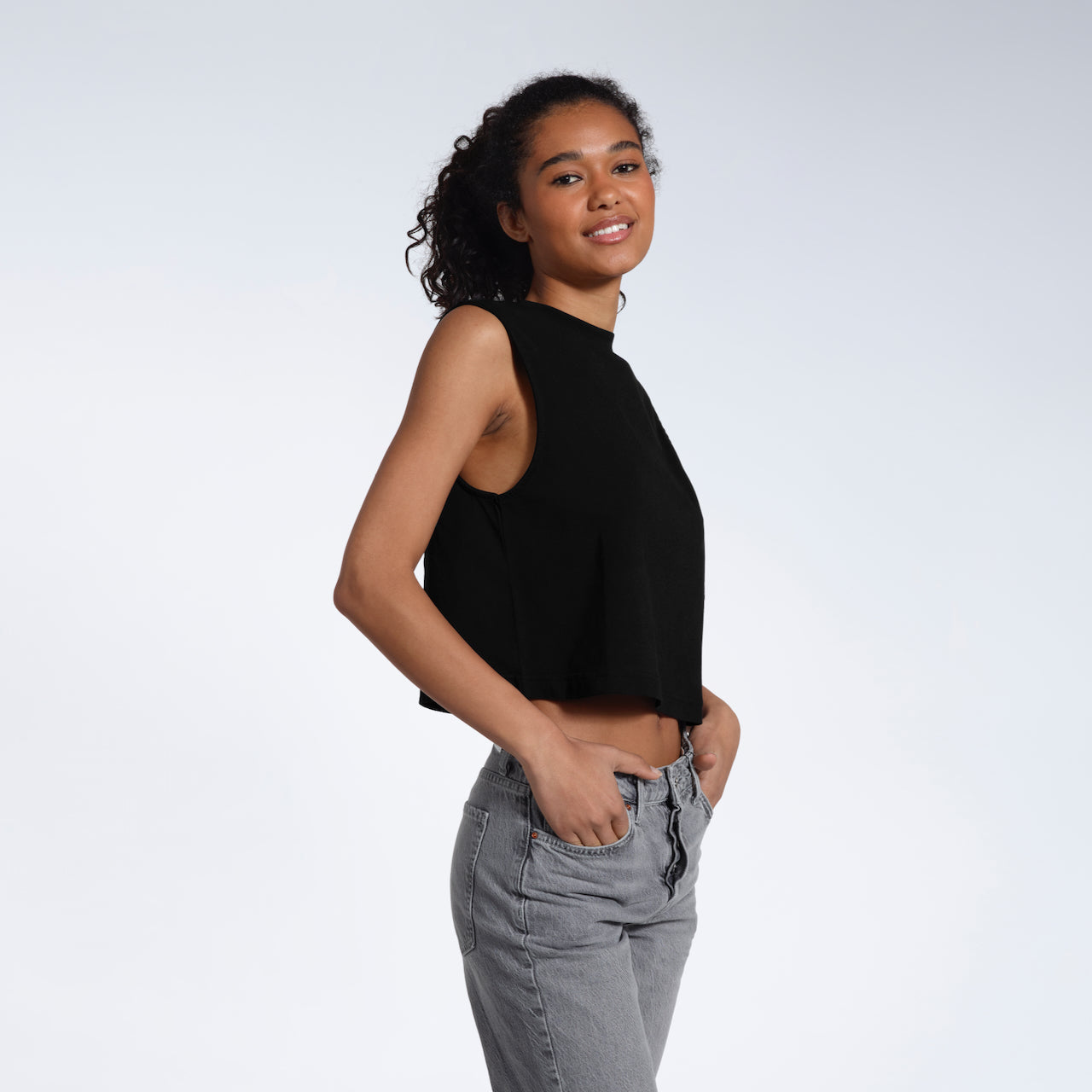 A woman smiling at the camera with her hands in her jeans pockets, modelling a plain black sleeveless organic cotton tank top.