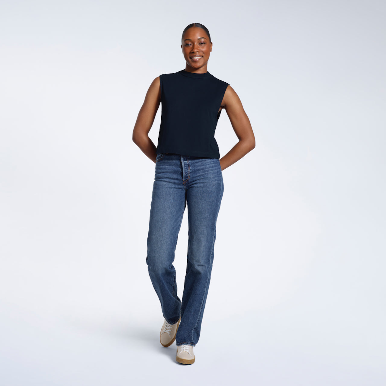 A full body photo of a woman smiling with her arms behind her back, modelling a plain navy sleeveless organic cotton tank top.
