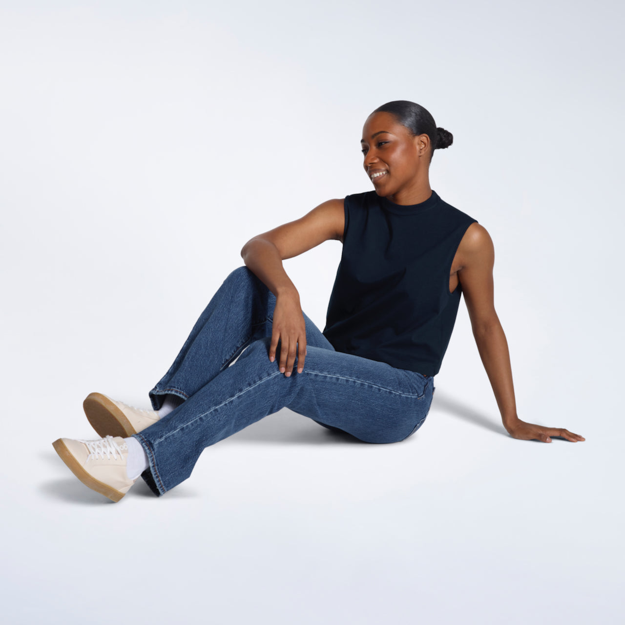 A woman sitting on the floor smiling, modelling a plain navy sleeveless organic cotton tank top.