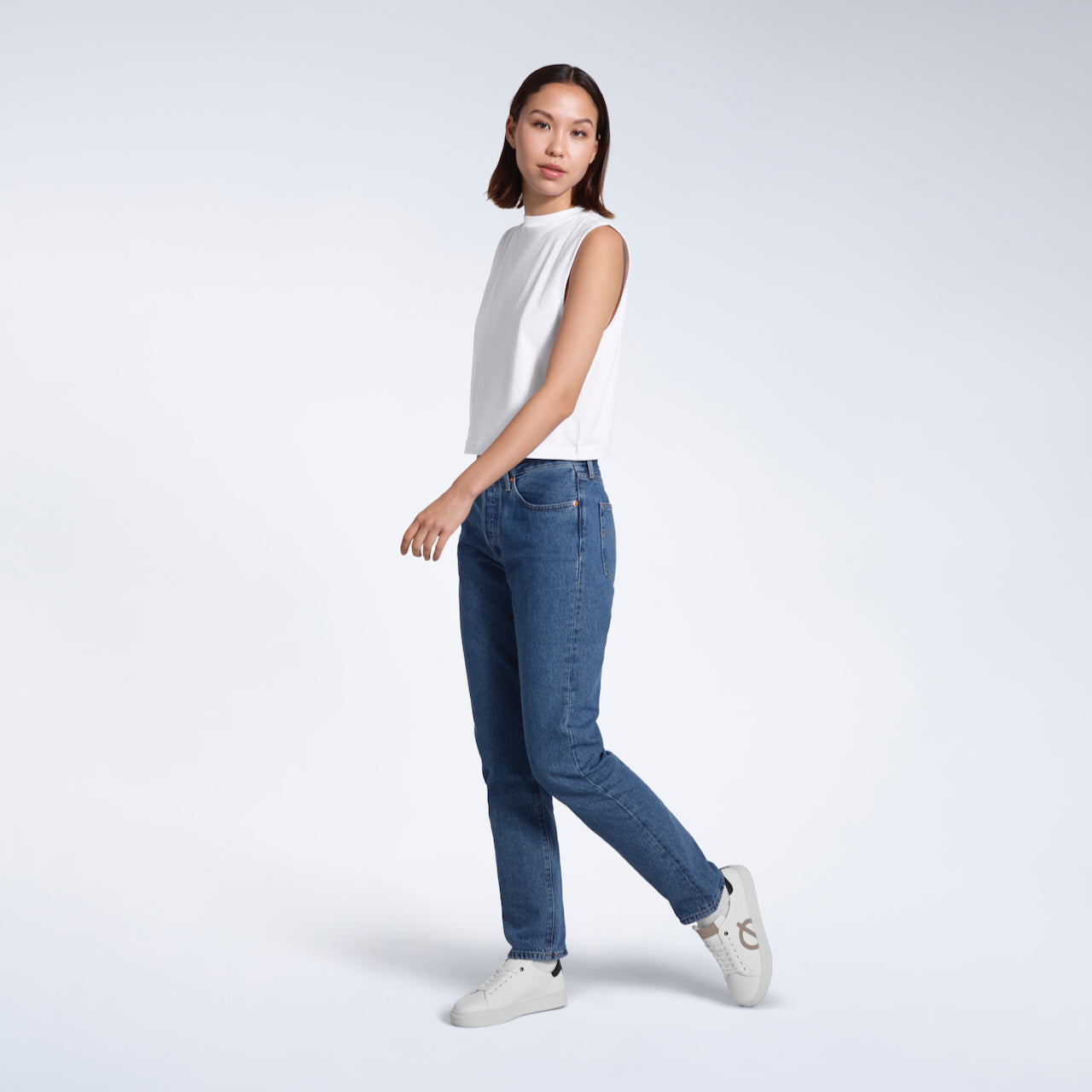 A full body photo of a woman walking, modelling a plain white sleeveless organic cotton tank top.