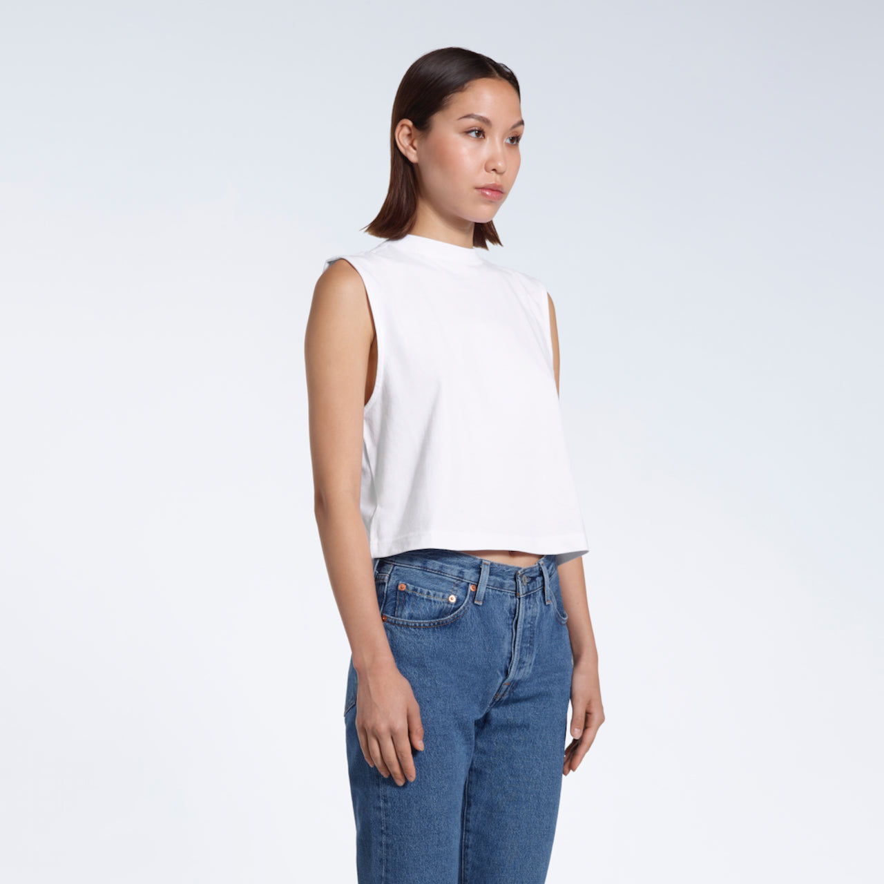A woman at an angle with her hands by her sides, modelling a plain white sleeveless organic cotton tank top.