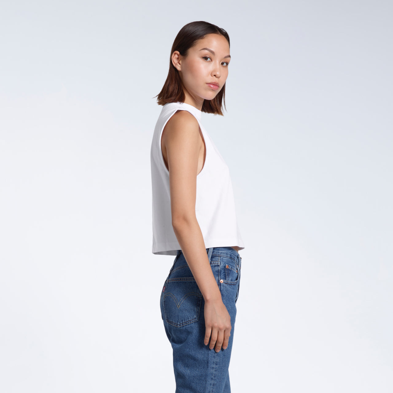 A woman facing side on and looking at the camera, modelling a plain white sleeveless organic cotton tank top.