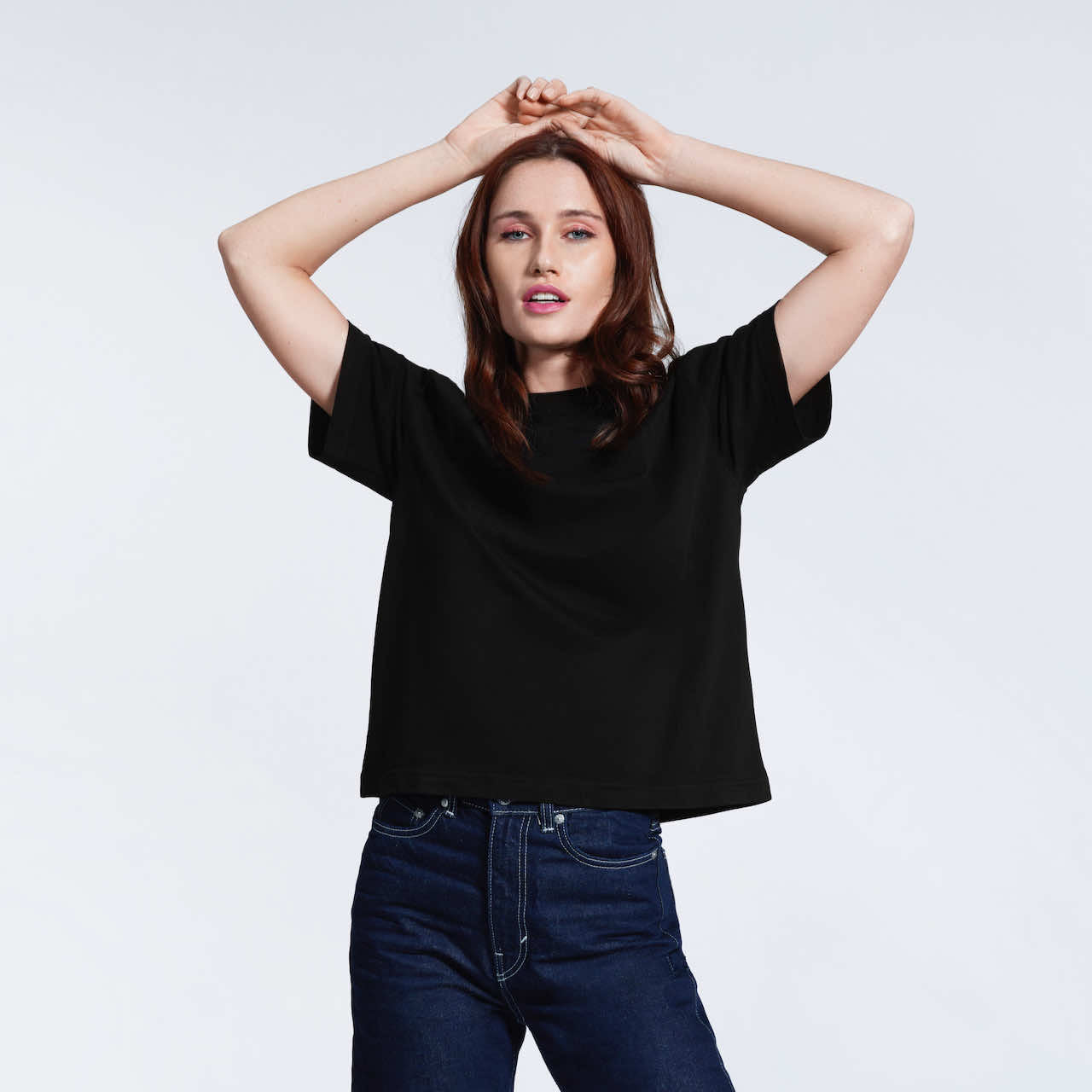 A woman with her hands on her head looking at the camera, modelling a plain black boxy, heavyweight organic cotton t shirt.
