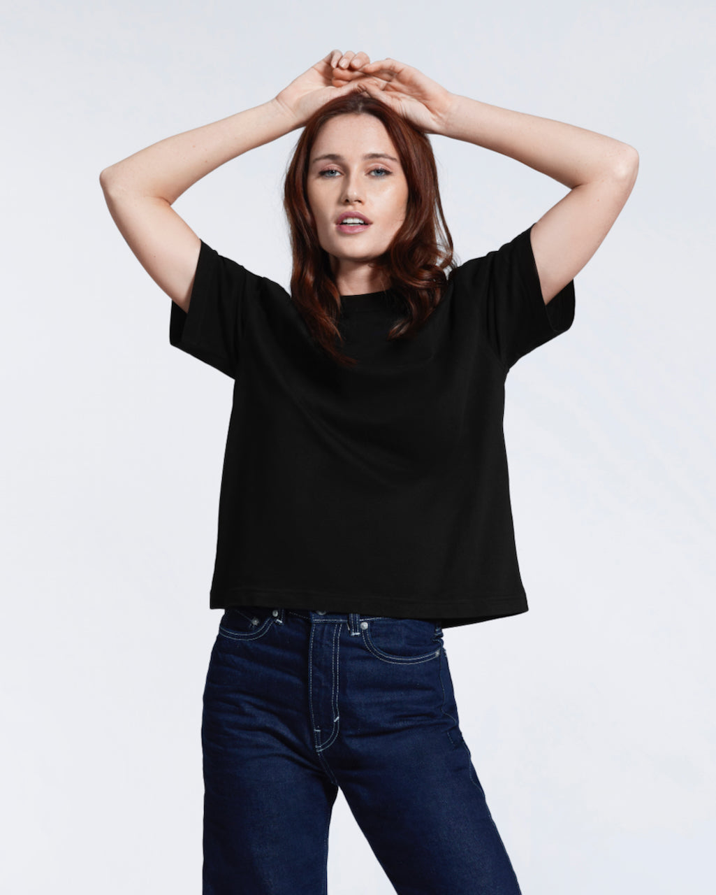 A woman with her hands on her head looking into the camera, modelling a plain black boxy, heavyweight organic cotton t shirt.
