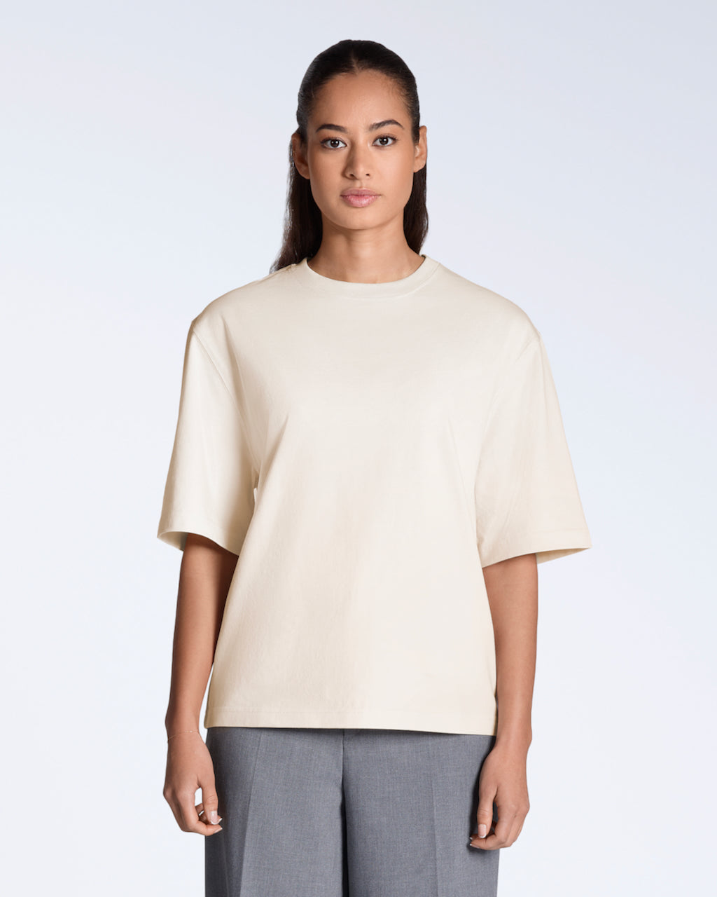 A woman face on looking into the camera, modelling a plain natural coloured, boxy, heavyweight, organic cotton t shirt.