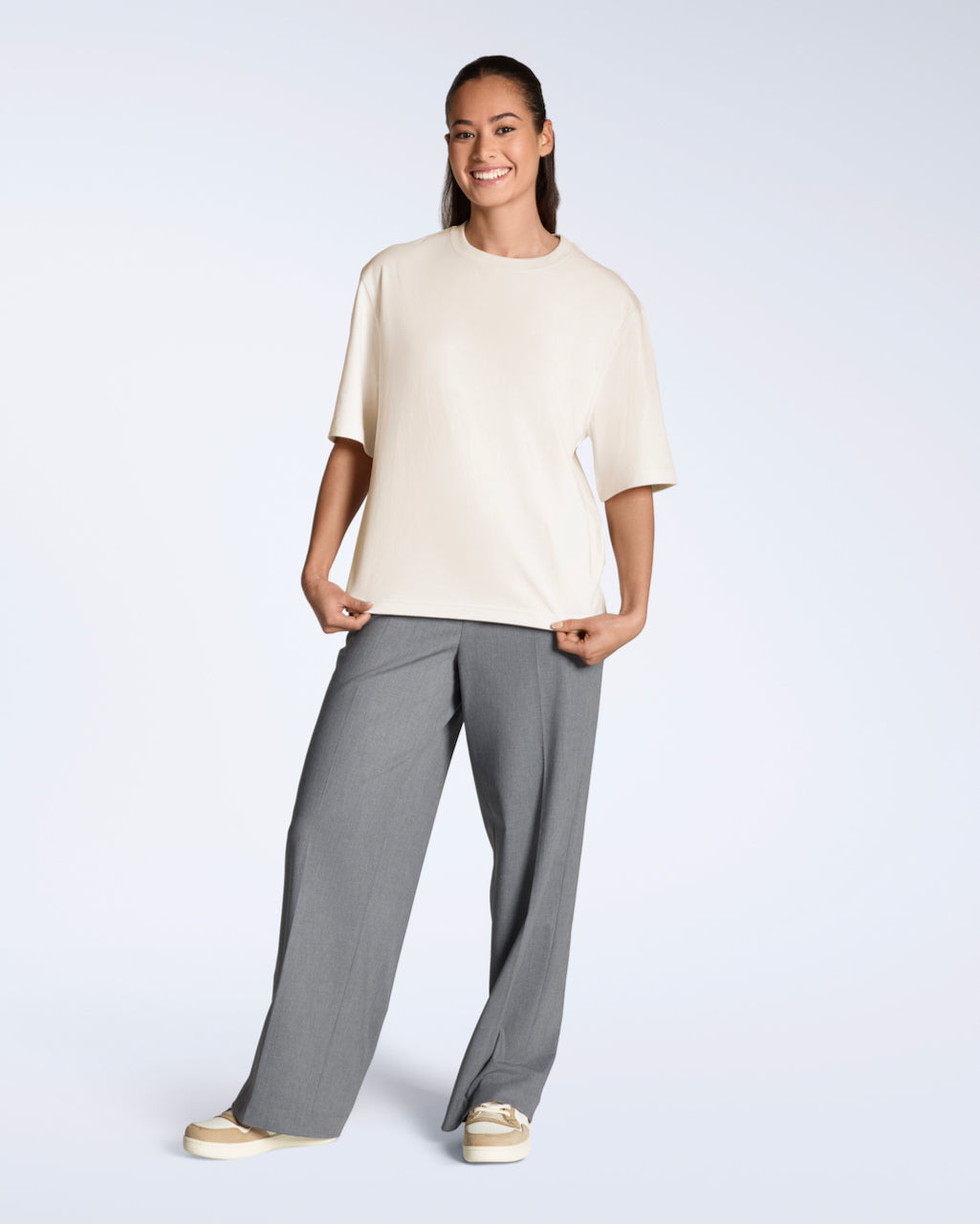A full body photo of a woman smiling into the camera, modelling a plain natural coloured, boxy, heavyweight, organic cotton t shirt.
