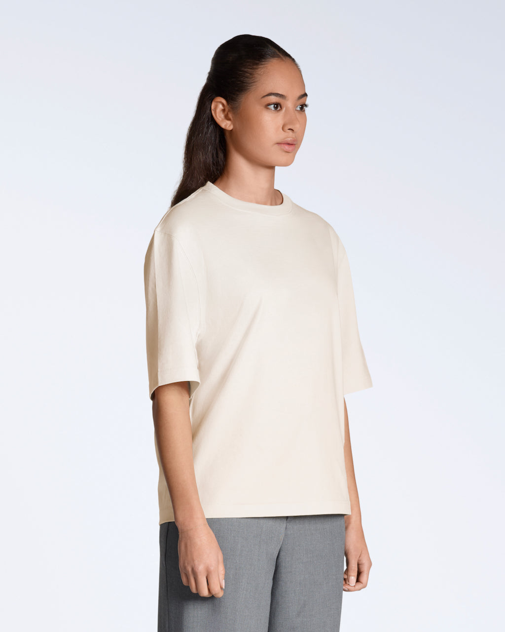 A woman at an angle to the camera with her arms by her side, modelling a plain natural coloured, boxy, heavyweight, organic cotton t shirt.