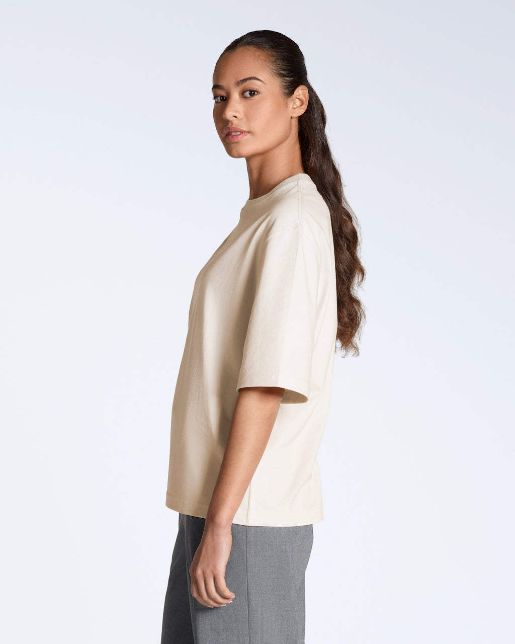 A woman facing side on looking back at the camera, modelling a plain natural coloured, boxy, heavyweight, organic cotton t shirt.