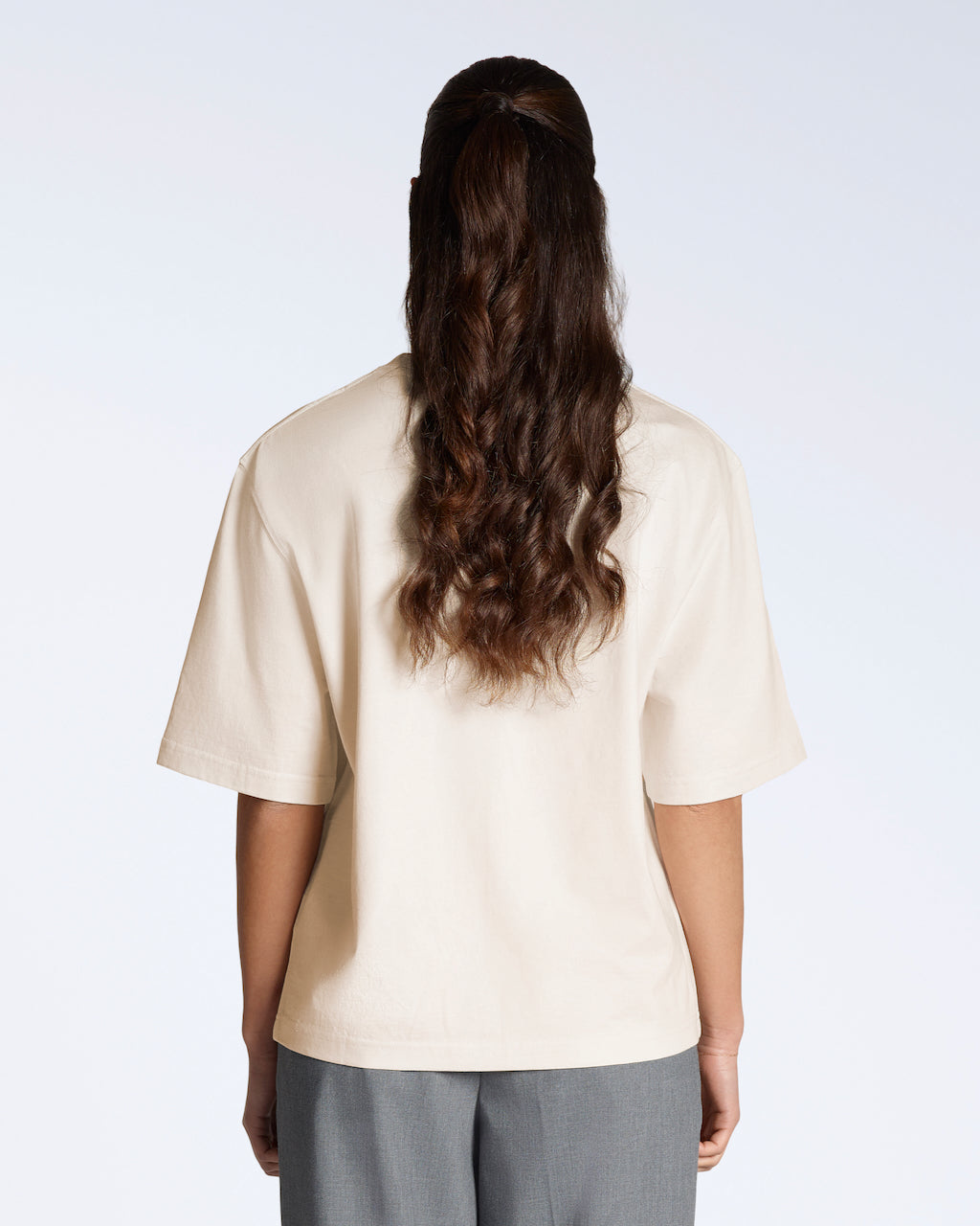 A woman facing away from the camera, modelling a plain natural coloured, boxy, heavyweight, organic cotton t shirt.