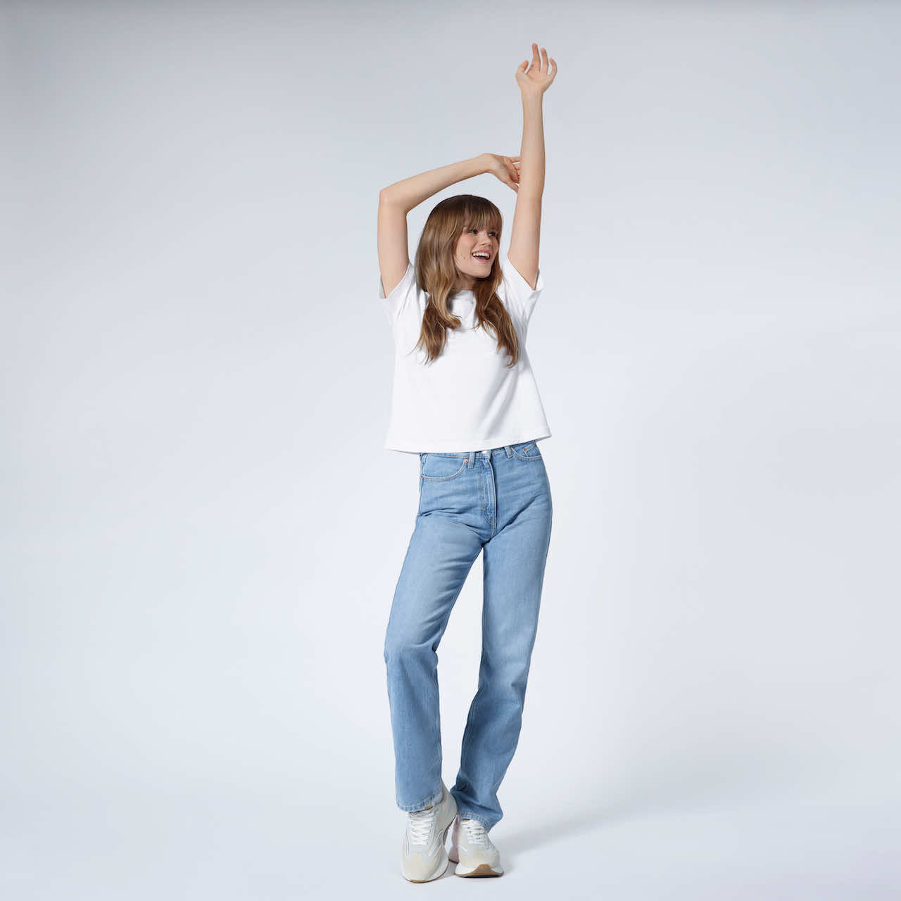 A full body photo of a woman with her arms above her head smiling, modelling a plain white boxy heavyweight organic cotton t shirt.