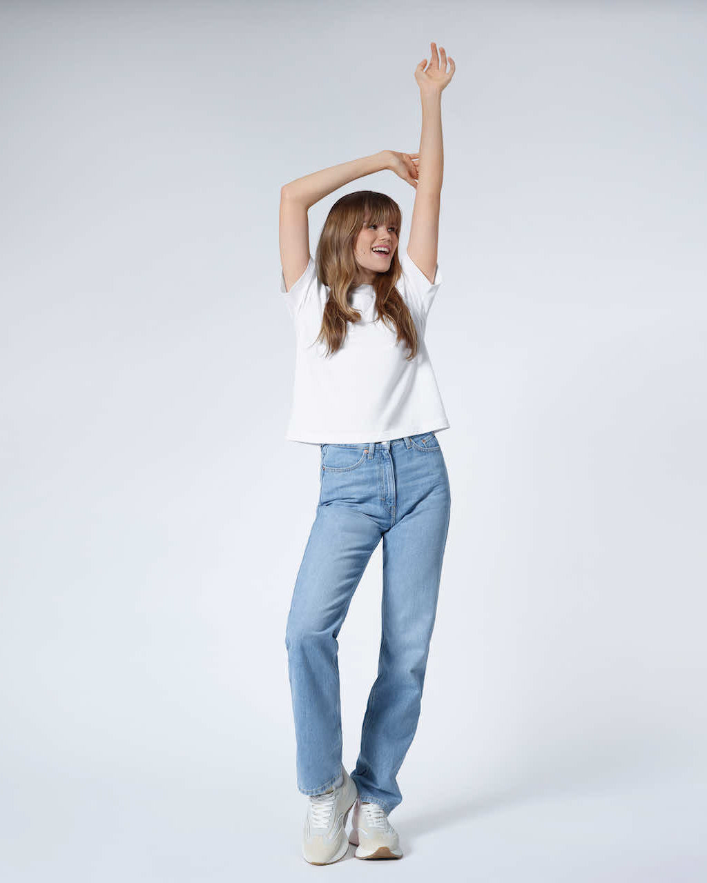 A full body photo of a woman with her arms above her head smiling, modelling a plain white boxy heavyweight organic cotton t shirt.