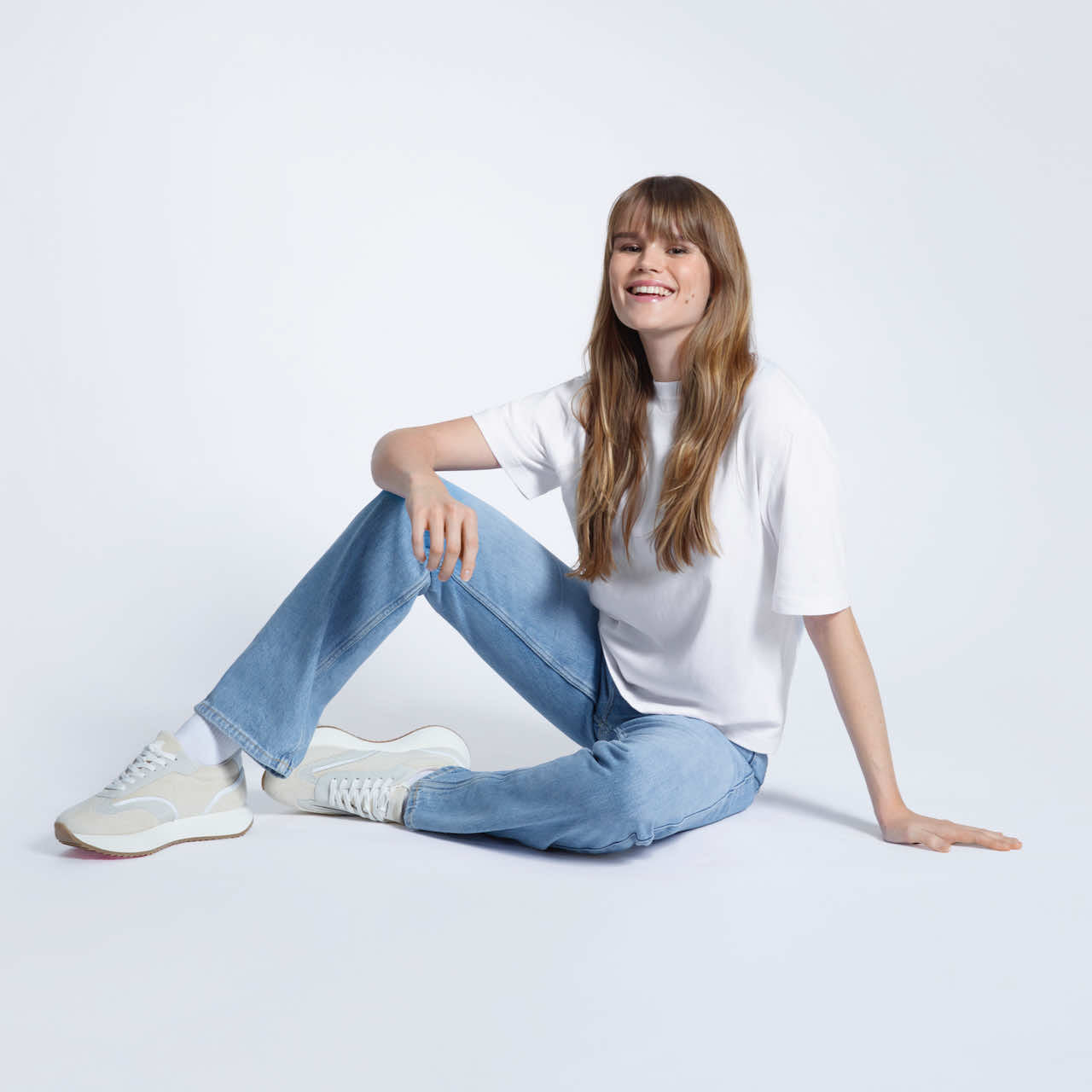 A woman sitting on the floor smiling, modelling a plain white, boxy, heavyweight organic cotton t shirt.