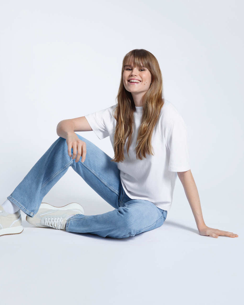 A woman sitting on the floor smiling, modelling a plain white, boxy, heavyweight organic cotton t shirt.
