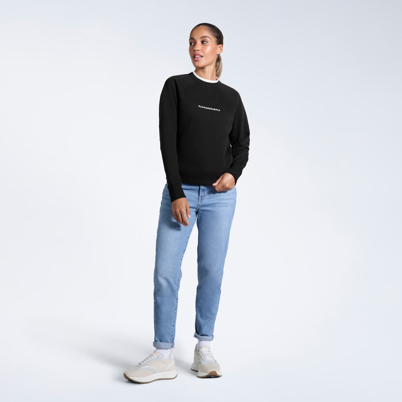 A full body photo of a woman looking away from the camera, modelling a branded black heavyweight organic cotton sweatshirt.