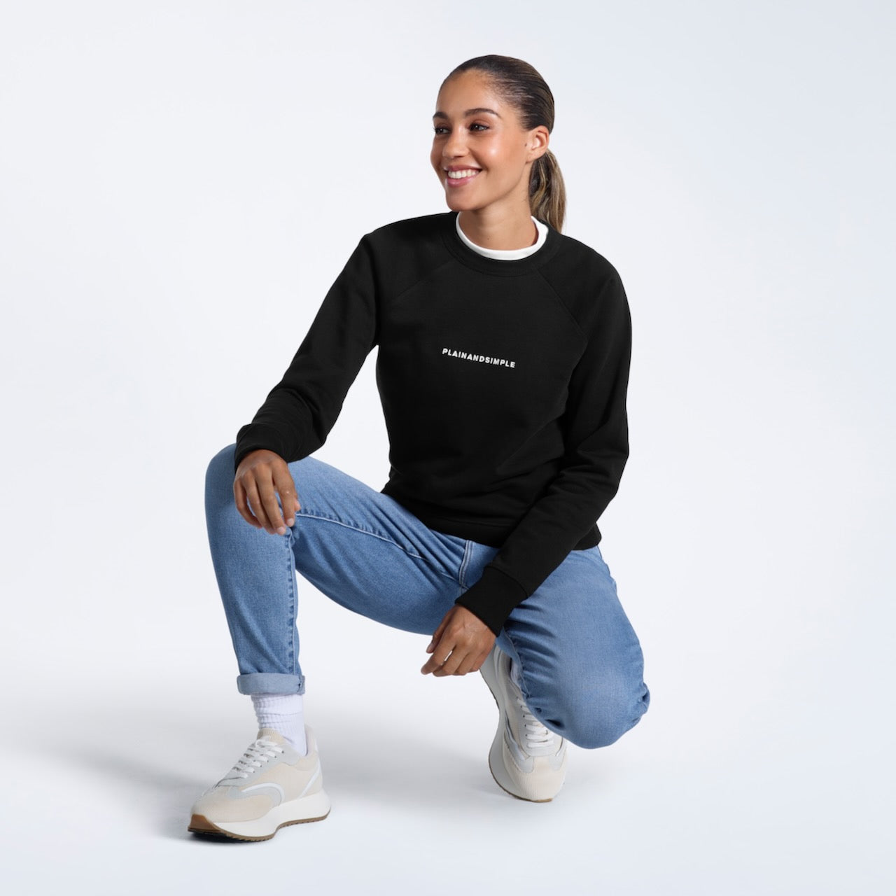 A woman crouching down smiling, modelling a black branded heavyweight organic cotton sweatshirt.