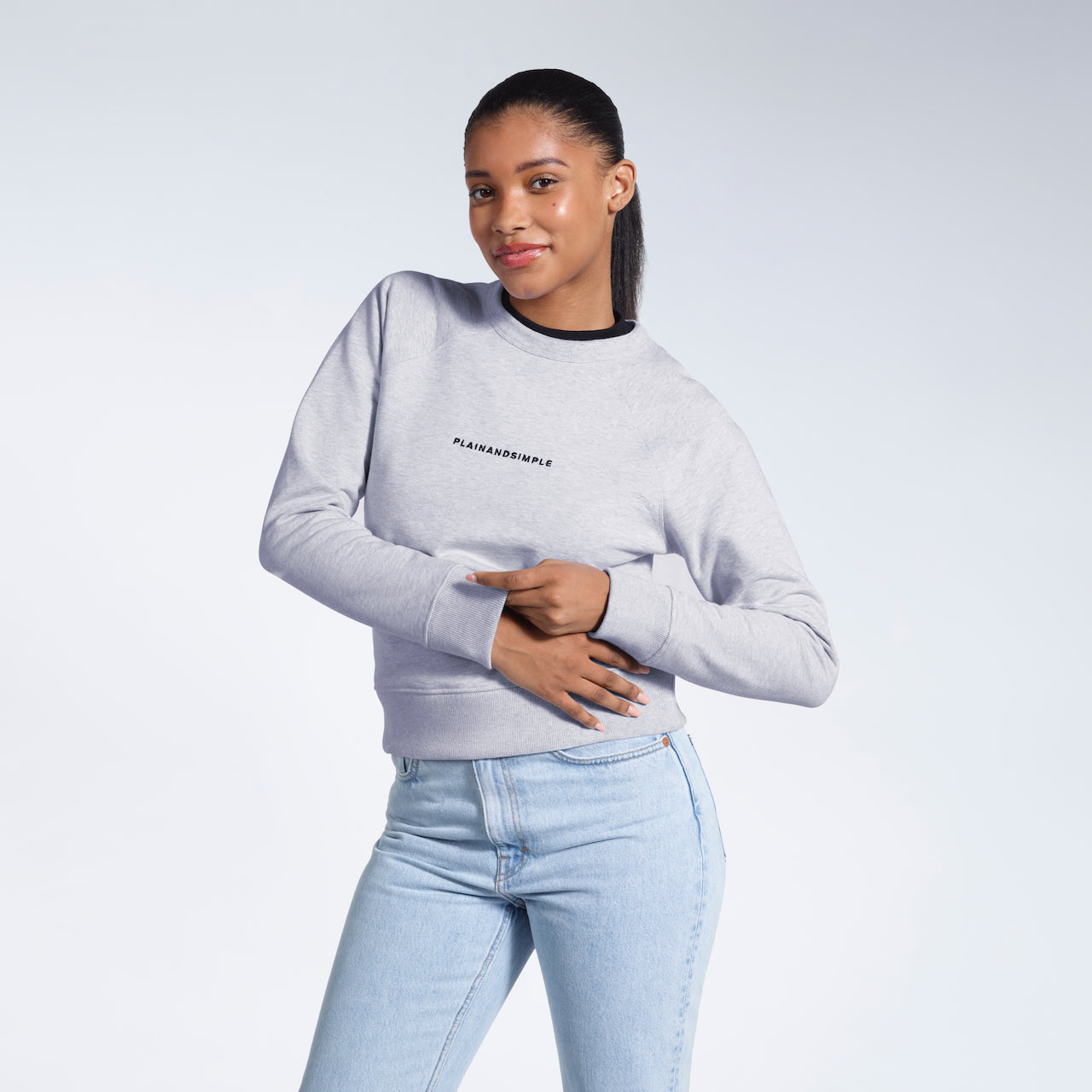 A woman smiling into the camera touching her hands, modelling a grey branded heavyweight organic cotton sweatshirt.