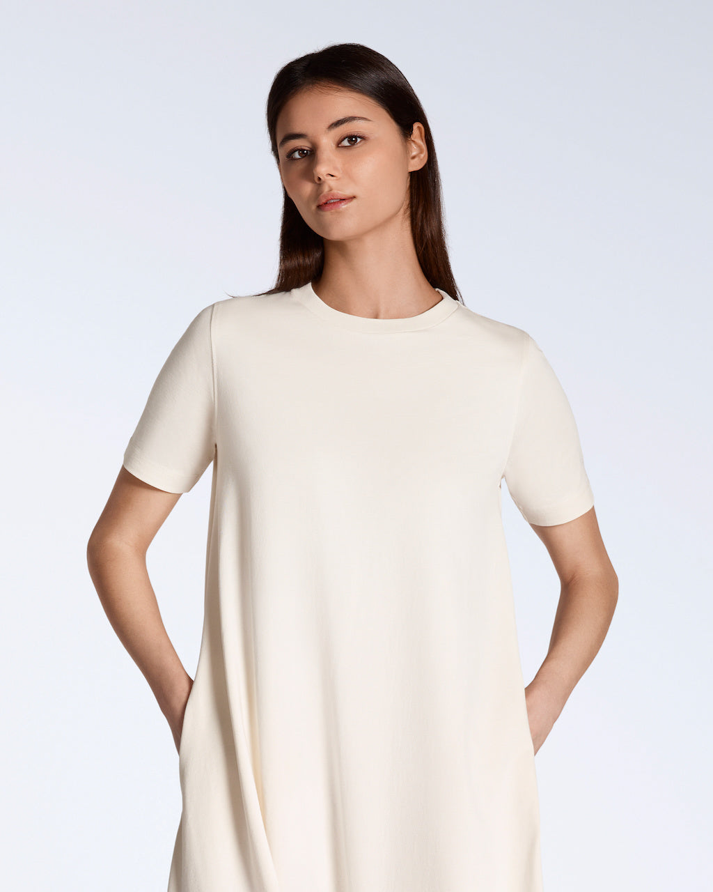 A woman looking into the camera with her hands in her pockets, modelling a plain natural coloured, long line, maxi organic cotton t shirt dress.