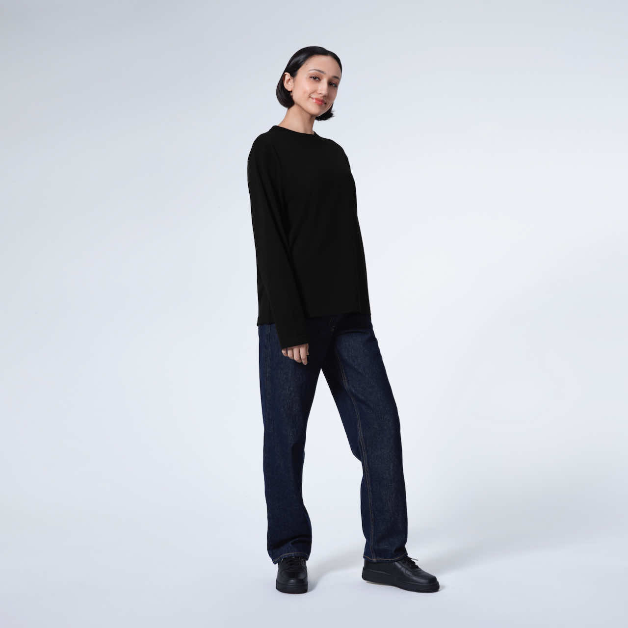 A full body photo of a woman looking into the camera smiling, modelling a plain black long sleeve organic cotton t shirt.