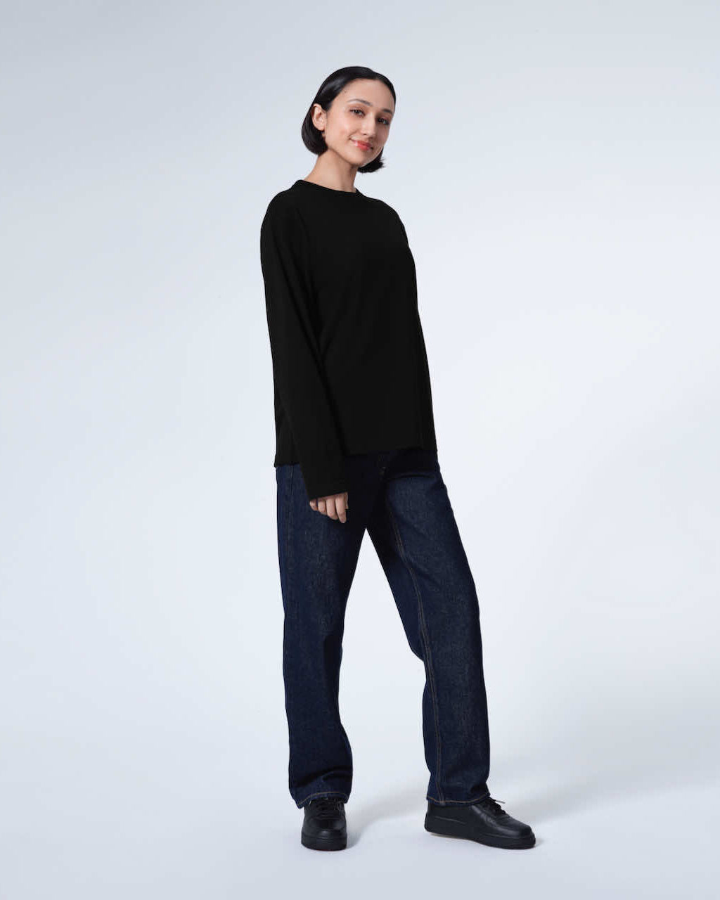A full body photo of a woman looking into the camera smiling, modelling a plain black long sleeve organic cotton t shirt.