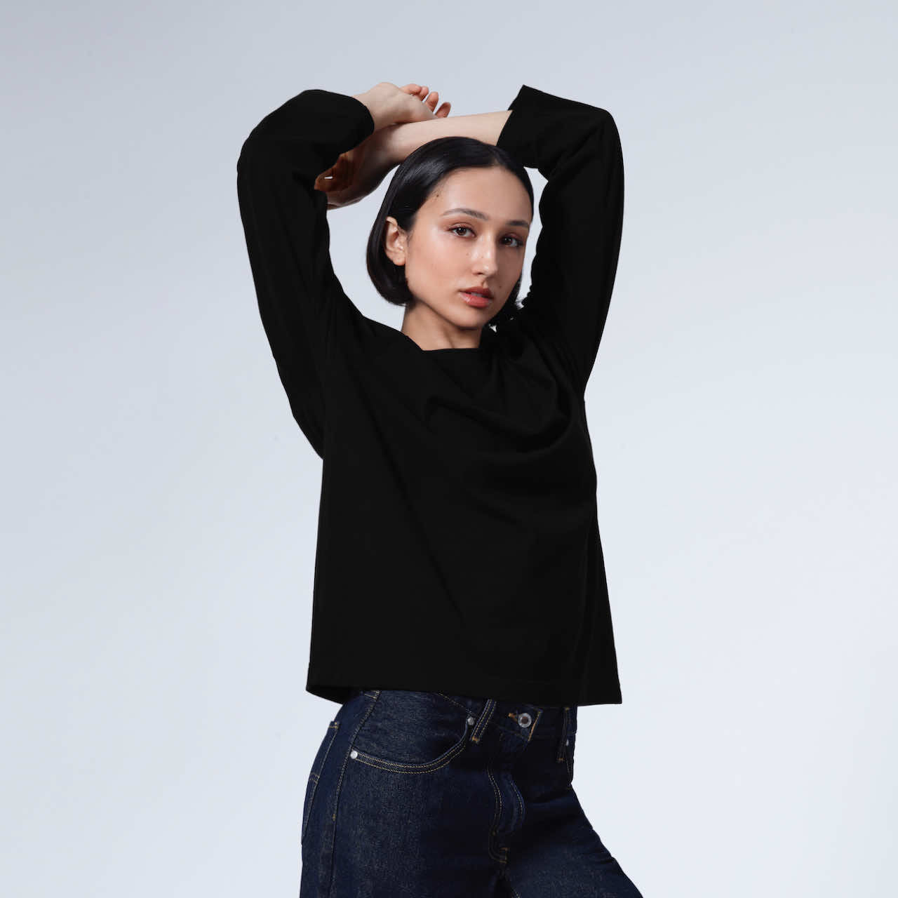 A woman looking into the camera with her arms above her head, modelling a plain black long sleeve organic cotton t shirt.