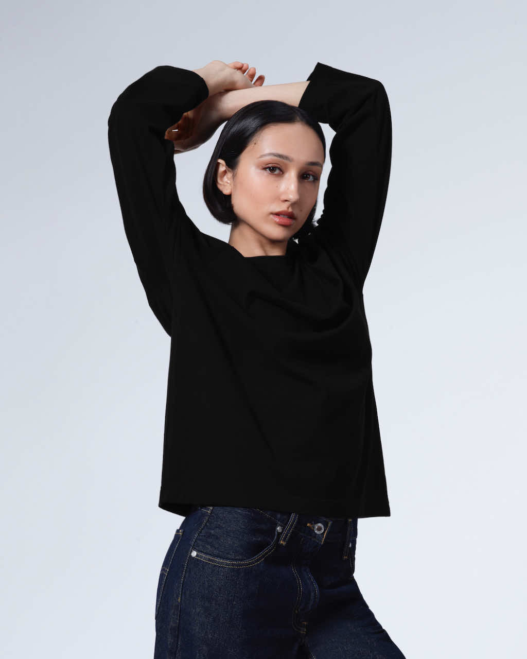A woman looking into the camera with her arms above her head, modelling a plain black long sleeve organic cotton t shirt.