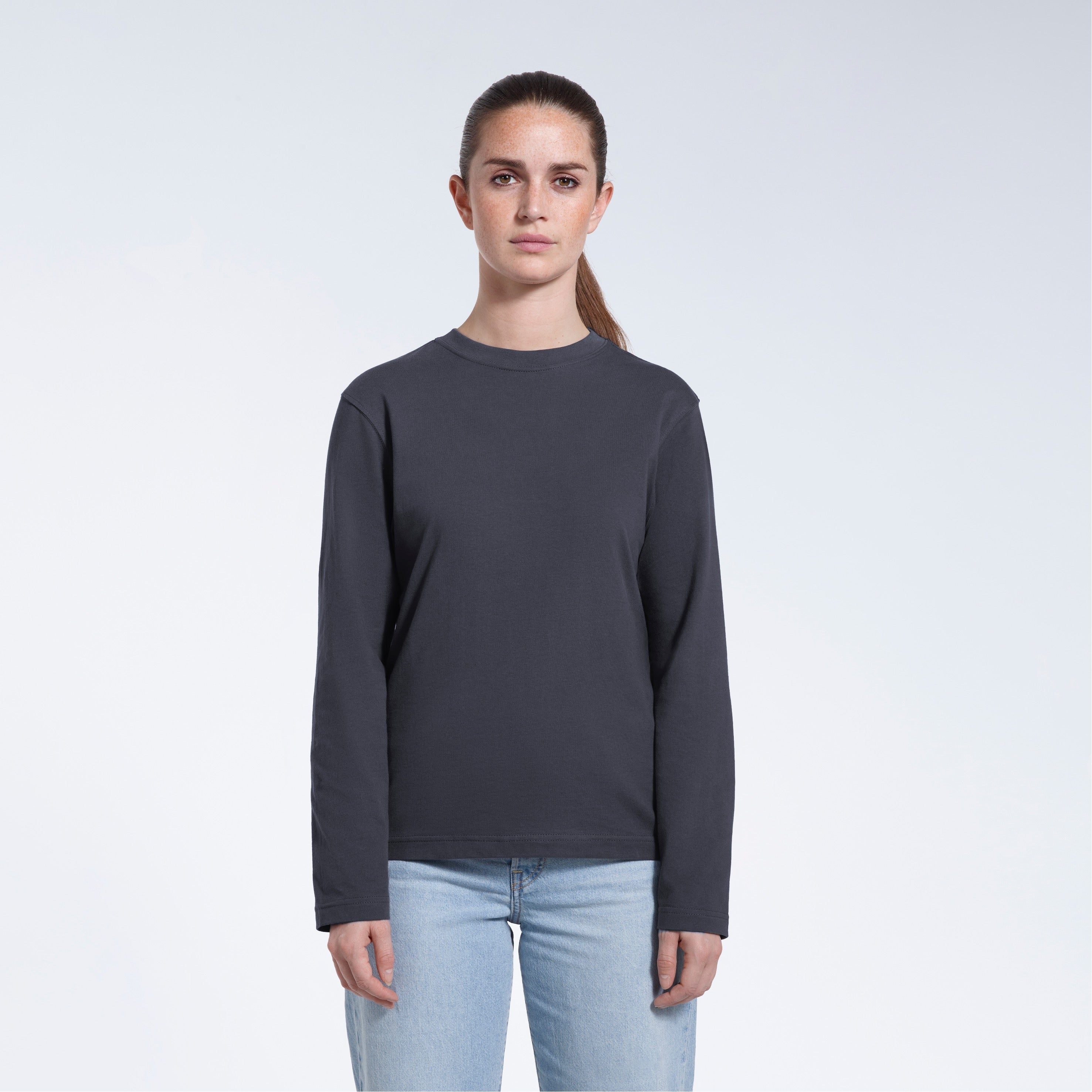 A woman standing front on with her arms by her sides, modelling a plain dark grey long sleeve organic cotton t shirt.