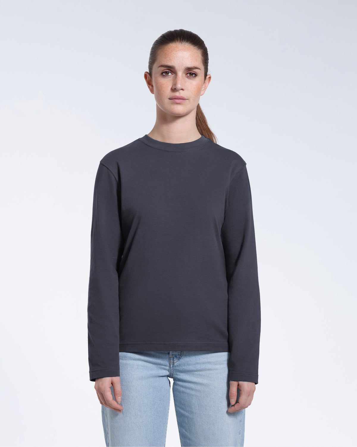A woman standing front on with her arms by her sides, modelling a plain dark grey long sleeve organic cotton t shirt.