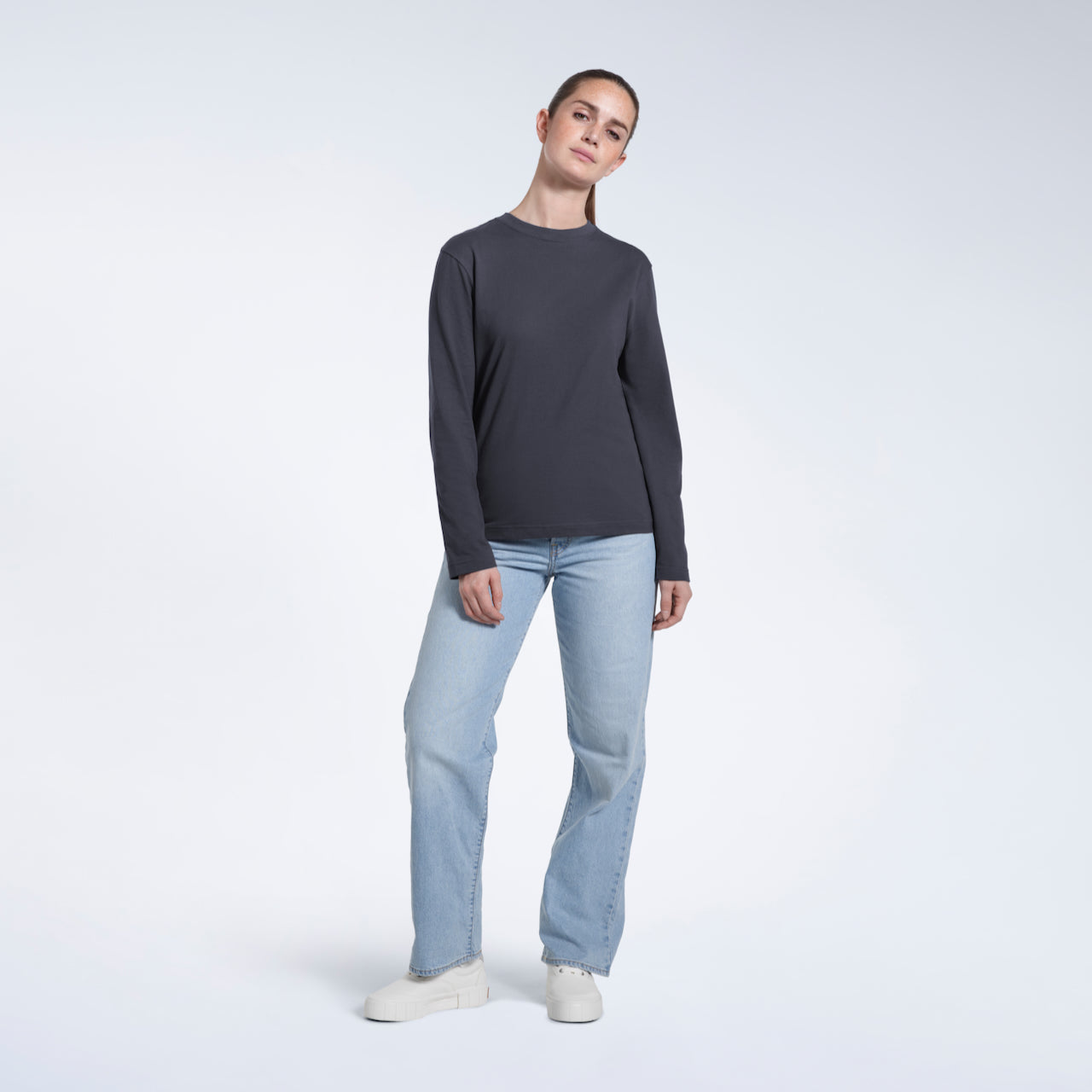 A full body photo of a woman looking into the camera, modelling a plain dark grey long sleeve organic cotton t shirt.