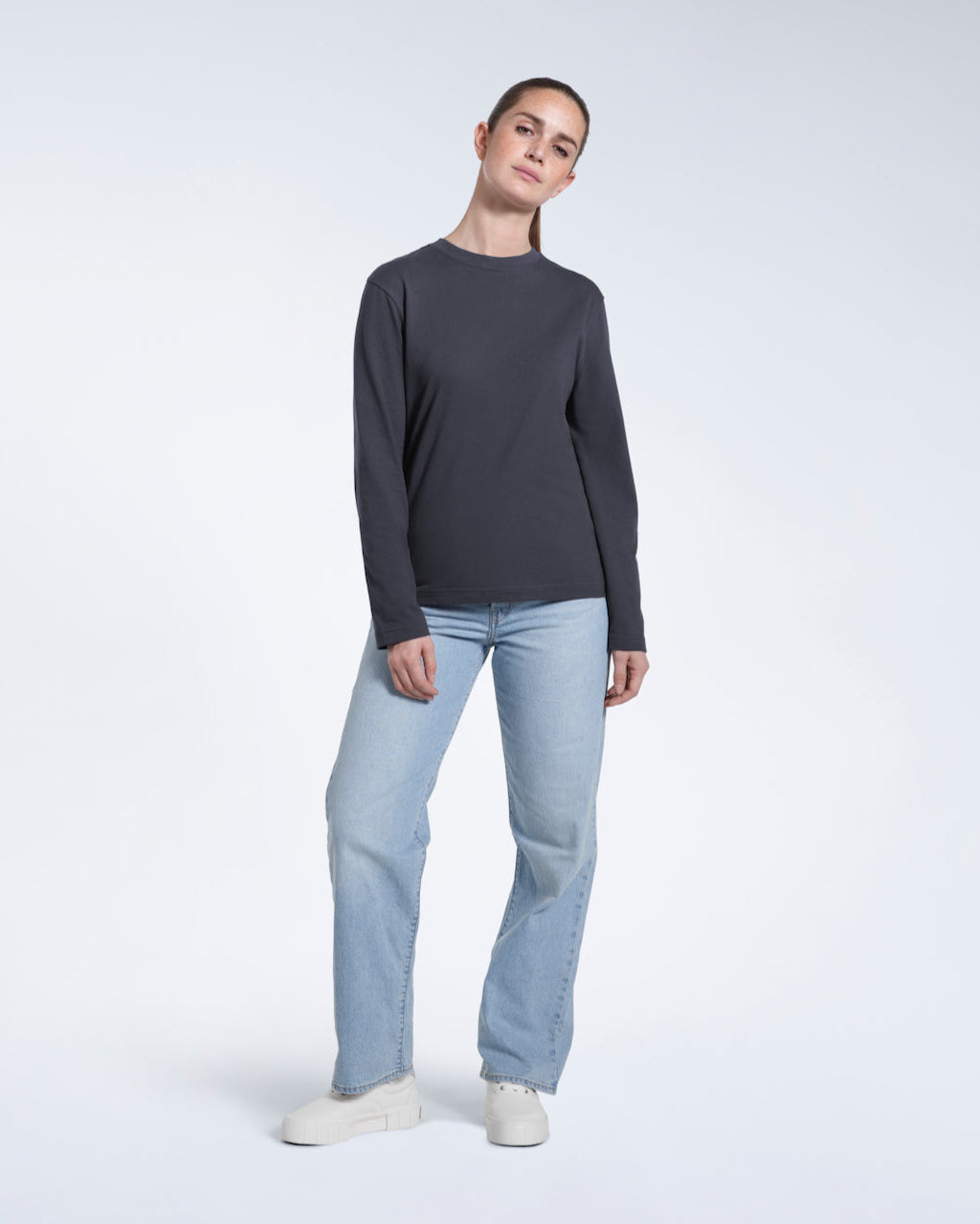 A full body photo of a woman looking into the camera, modelling a plain dark grey long sleeve organic cotton t shirt.