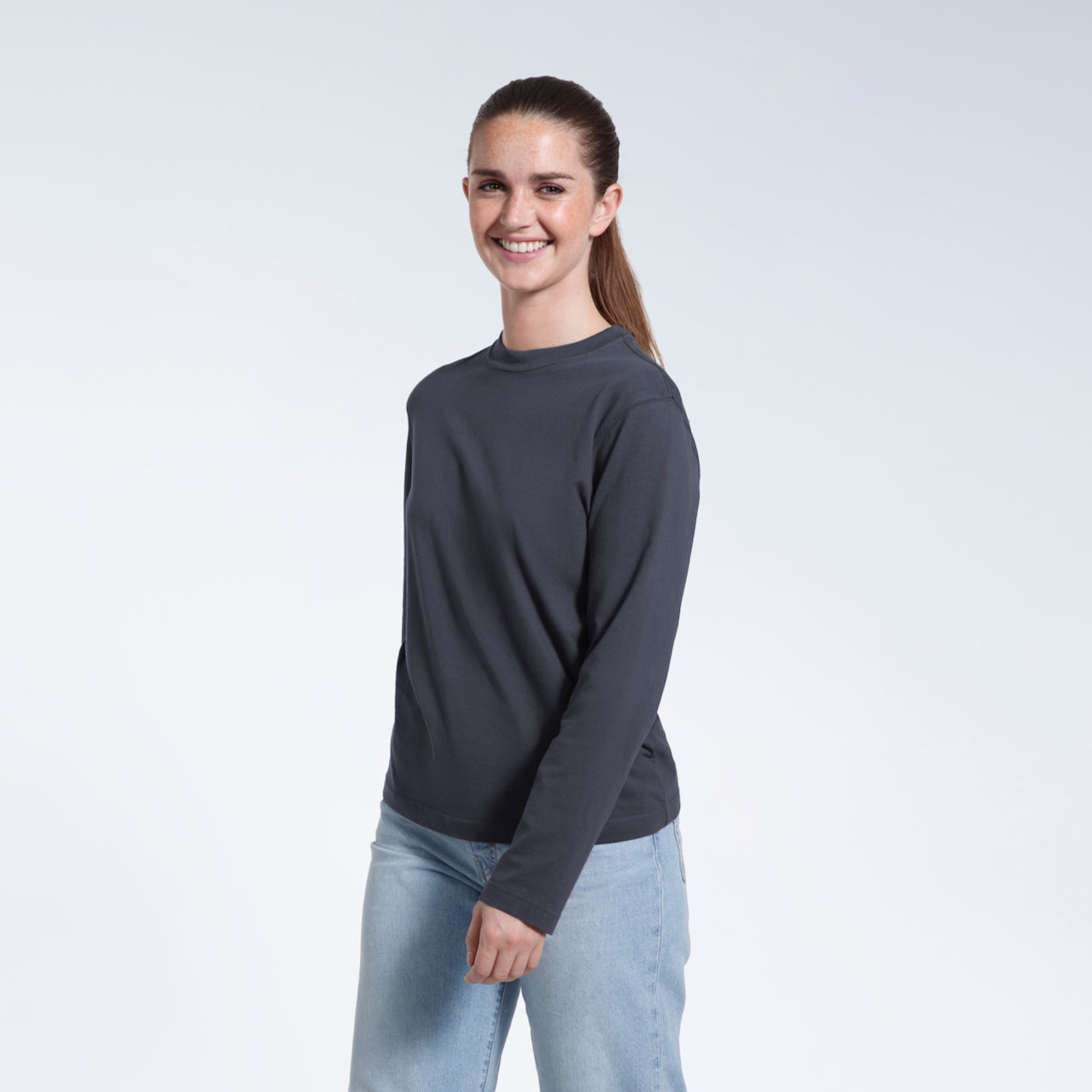 A woman smiling into the camera, modelling a plain dark grey long sleeve organic cotton t shirt.