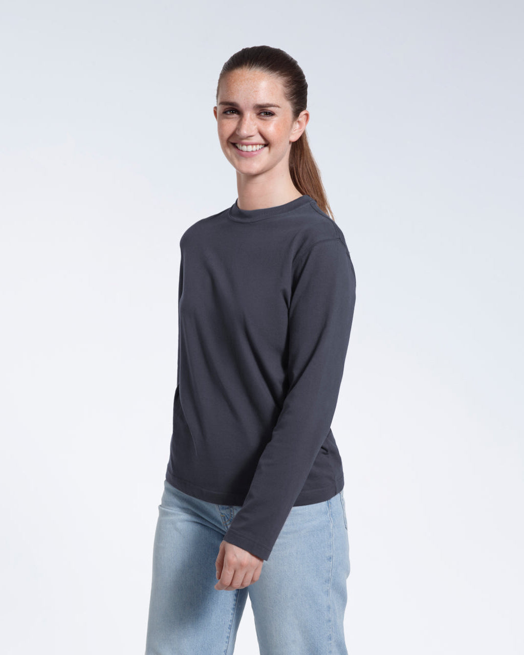 A woman smiling into the camera, modelling a plain dark grey long sleeve organic cotton t shirt.
